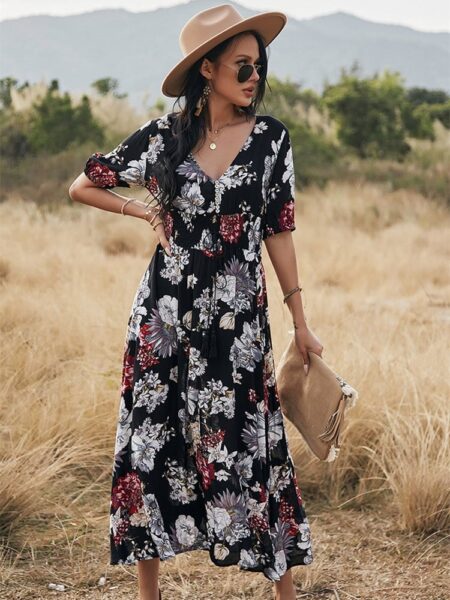 Femme dans un champs de blé portant une robe noire longue à fleurs avec u, chapeau sur la tête et une pochette dans sa main droite