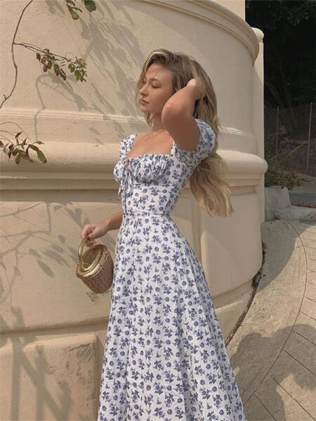 Femme blonde portant une robe longue blanche avec un imprimé fleuri bleu. Elle passe sa main gauche dans ses cheveux et dans sa main droite, elle tient un petit sac