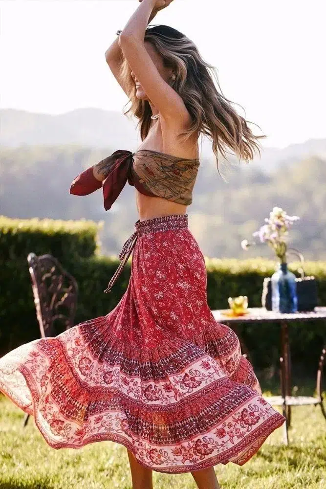 Femme typé bohème portant une jupe longue et ample rouge et fleurie avec un foulard autour de sa poitrine. Elle est dans un jardin et elle tourbillonne sur elle-même
