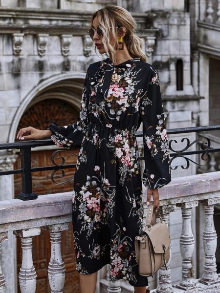 Robe longue fleurie élégante à manches longues de couleur noire, portée par une femme debout en ville portant un sac à main beige